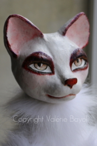 Wachsame Katzenfigur mit prüfendem Blick — Figurenkunst von Valérie Bayol Nahaufnahme des intensiv durchdringenden Blicks einer weißen Katze mit markantem braun-schwarzem Augenring von Bildhauerin Valérie Bayol aus eigener Holzmasse.